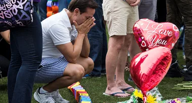 A man kneels at a memorial with "I Love You" heart balloons and flowers, appearing emotional with hands on his face.