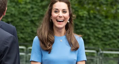 A woman with long brown hair smiles brightly, wearing a light blue dress with a necklace, standing outdoors.