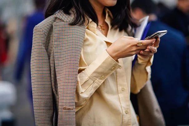 Person in a yellow shirt with a checkered jacket draped over shoulders, typing on a smartphone with other people in blurry background.