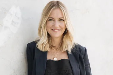 Blonde woman with medium-length hair wearing a black blazer and layered necklaces, smiling against a light-colored background.