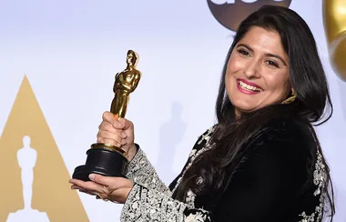 Woman smiling and holding an Oscar statuette, wearing ornate black and silver attire.