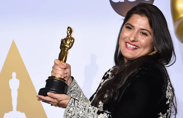 Woman smiling and holding an Oscar statuette, wearing ornate black and silver attire.