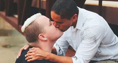 Two people sharing a kiss, sitting on a bench. One has short blond hair, the other has short dark hair.
