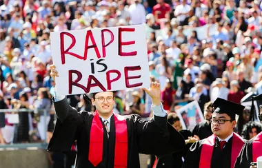 Graduate holds "Rape is Rape" sign at a commencement ceremony, standing among fellow graduates in caps and gowns.
