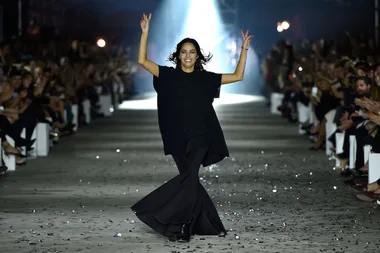 Person dressed in black walking down a runway, arms raised, with audience clapping on either side.