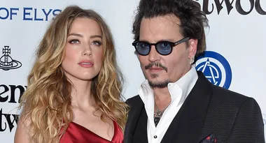 A woman in a red dress and a man in black suit with sunglasses pose together at a formal event.