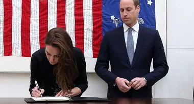 Kate Middleton signing a book as Prince William stands by, with a red, white, and blue quilt in the background.