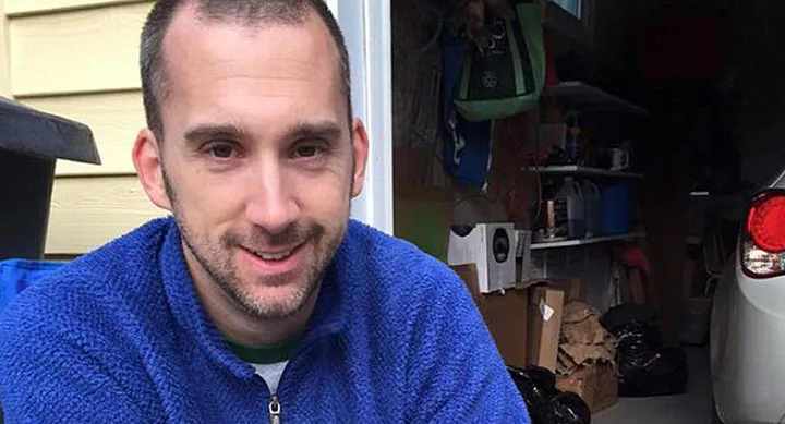 Man in a blue fleece jacket sitting in a garage with shelves and a car visible in the background.