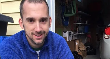 Man in a blue fleece jacket sitting in a garage with shelves and a car visible in the background.
