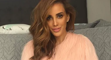 Woman with long wavy hair wearing a pink fur coat seated on a gray bed.