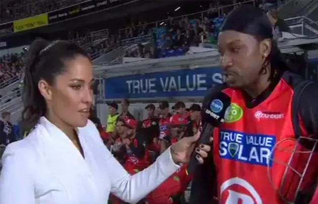 An interviewer in white speaks to a cricketer in a red uniform holding a microphone in a stadium.