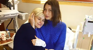 Two women embracing in a hospital room, one wearing a blue sweater. Medical equipment is visible in the background.
