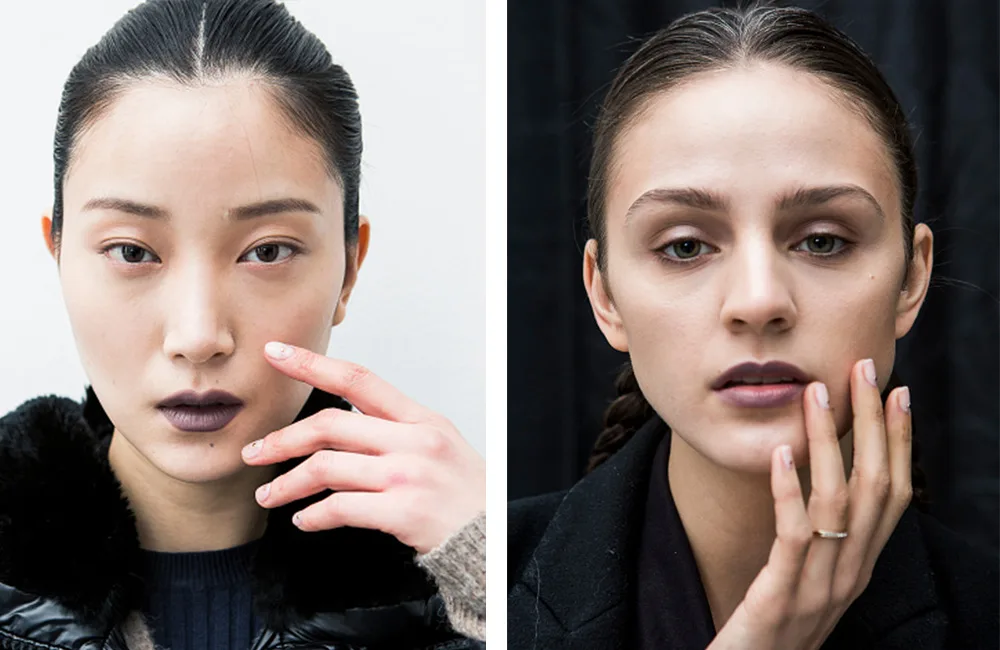 Two women with neutral expressions and dark lipstick, each touching their face with a hand, against a neutral background.