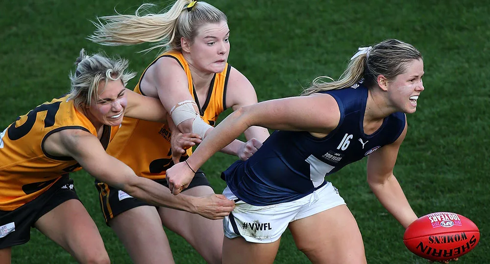 Two female Australian rules football players in action; blue team member holding the ball, yellow team members attempting a tackle.