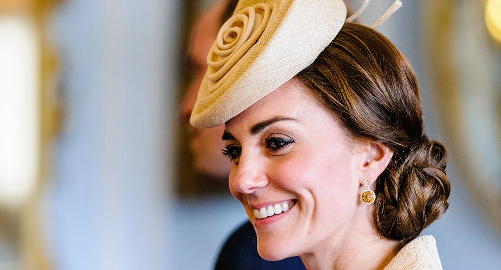 A woman smiling, wearing a beige hat with a rose pattern and gold earrings, with brown hair styled in an updo.