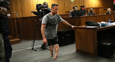 Man walking without prosthetics in a courtroom, cameraman in background, officials seated at a desk.