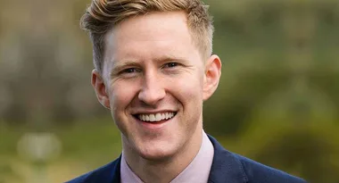A man in a suit smiling outdoors with blurred greenery in the background.