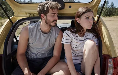 Two people sitting in the open trunk of a yellow car, outdoors on a sunny day.