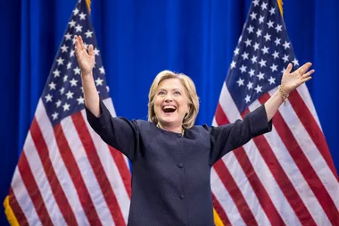 A person in a dark suit with arms raised, smiling, stands in front of two US flags.