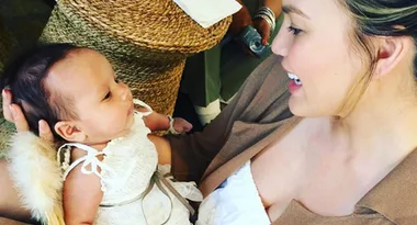A woman in a brown jacket holds a smiling baby dressed in a white outfit, in a cozy setting.