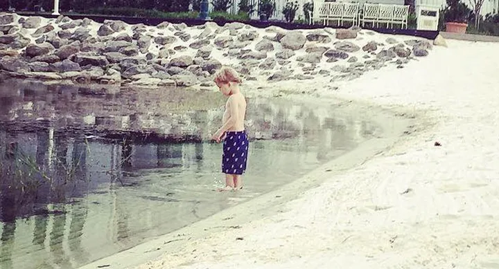 A child wearing dark shorts stands at the edge of a sandy shore looking at the water.