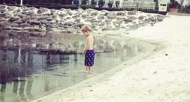 A child wearing dark shorts stands at the edge of a sandy shore looking at the water.