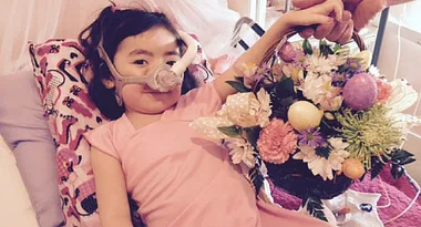 Young girl in a pink dress, wearing a nasal mask, holds a colorful flower basket while lying on a bed with a patterned pillow.