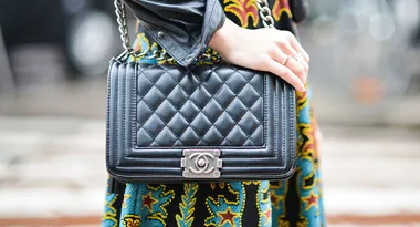 Quilted black Chanel handbag with a silver logo clasp, worn by a person in a colorful dress and leather jacket.