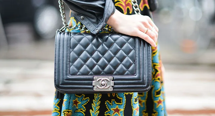 Quilted black Chanel handbag with a silver logo clasp, worn by a person in a colorful dress and leather jacket.