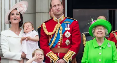 Royal family on a balcony, smiling and looking upwards. Woman in white holds a child, man in red uniform, elder woman in green hat.