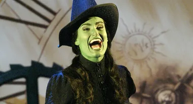 Green-skinned witch in black hat and outfit smiling broadly on stage.