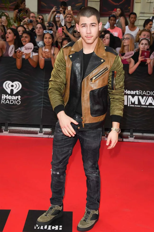 Man in a brown and green leather jacket on the red carpet with fans in the background at an iHeartRadio event.
