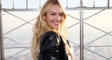 Blonde woman smiles near a cityscape from a high observation deck, wearing a black jacket.