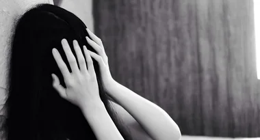 A person with long dark hair covers their face with hands, leaning against a wall, in a dimly lit room.