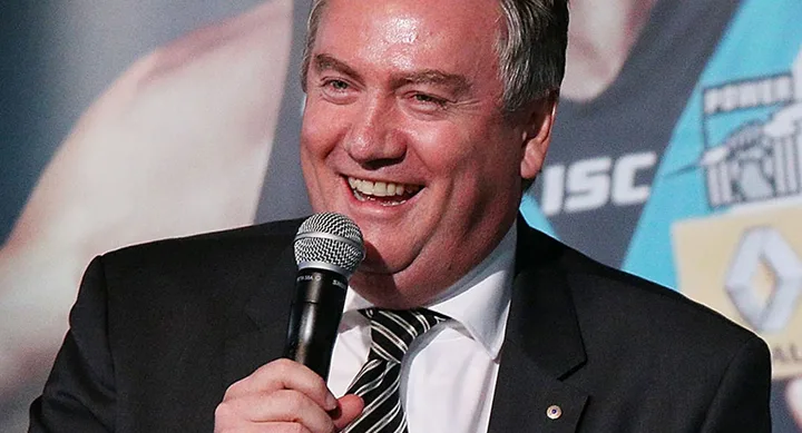 Man in a suit holding a microphone and smiling, with a sports poster in the background.