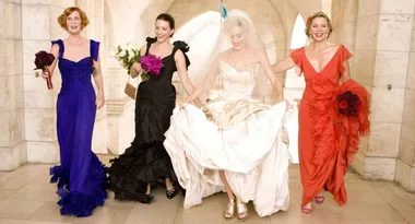 Bridesmaids in colorful dresses walk with a bride in a gown holding her train indoors.
