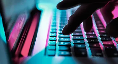 A hand typing on a laptop keyboard with blue and pink lighting.