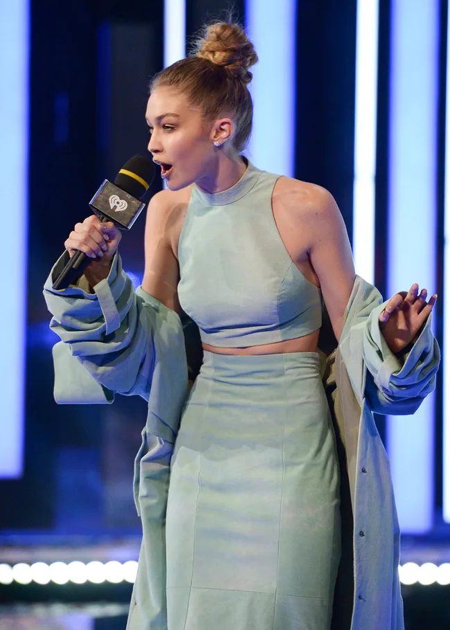 Model in light blue outfit speaking into a microphone on stage with bright vertical lights in the background.