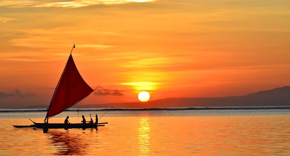 Sailboat with red sail and three people silhouetted against a vibrant orange sunset over calm water.