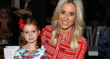 Two people smiling for a photo at an event; one adult with long blond hair and one child with a red bow in her hair.