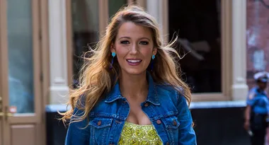 A woman with long hair in a denim jacket and yellow dress, smiling outdoors.