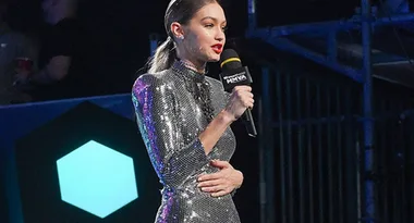 Woman in a silver sequin dress speaks into a microphone on stage at an event.