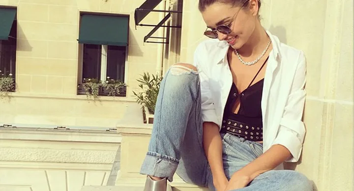 Woman in sunglasses and casual outfit sitting on a ledge, smiling in sunlight.