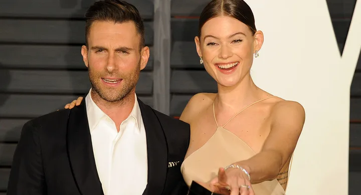 A smiling couple poses together; the woman shows her ring while the man has his arm around her at a formal event.