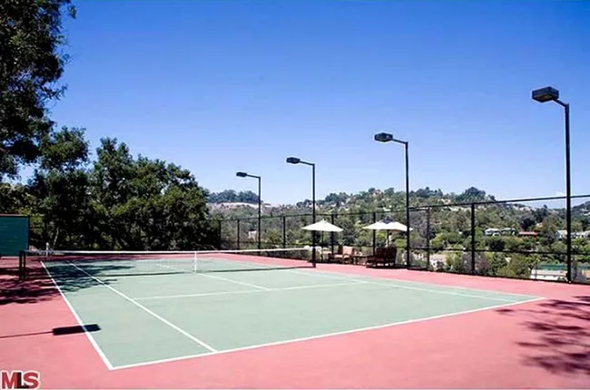 Tennis court with red surface, surrounded by trees with a clear blue sky and distant hills in the background.