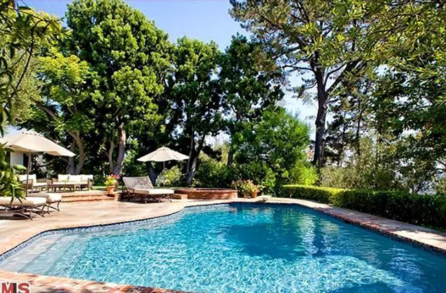 Outdoor pool surrounded by trees, lounge chairs, and umbrellas on a sunny day.