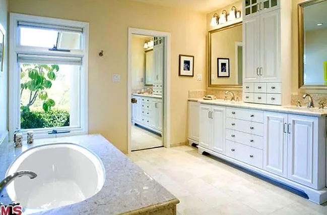 Elegant bathroom with a marble tub, large mirrors, and white cabinetry, featuring a window overlooking greenery.