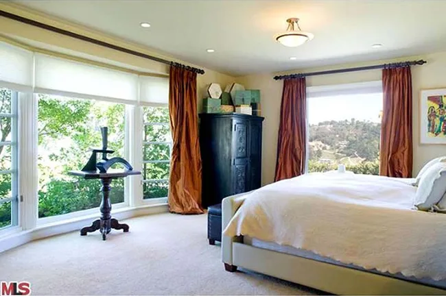 Bedroom with large windows, white bedding, brown curtains, dark cabinet, a table, and scenic outdoor view.