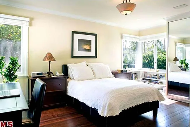 Cozy bedroom with a neatly made bed, desk with laptop, wooden flooring, large window, and mirrored closet door.
