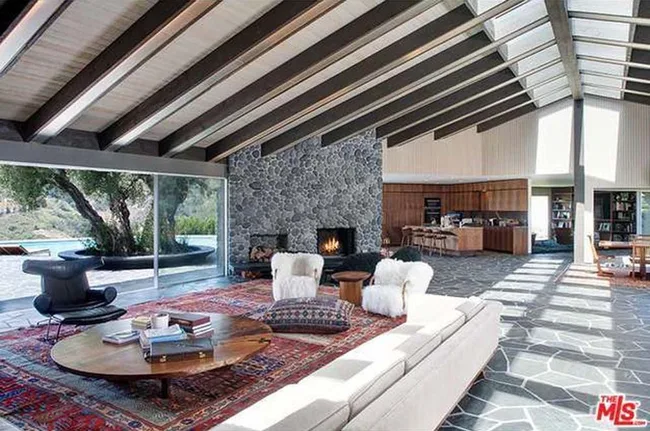A spacious living room with a stone fireplace, large windows, exposed beams, and modern furniture.